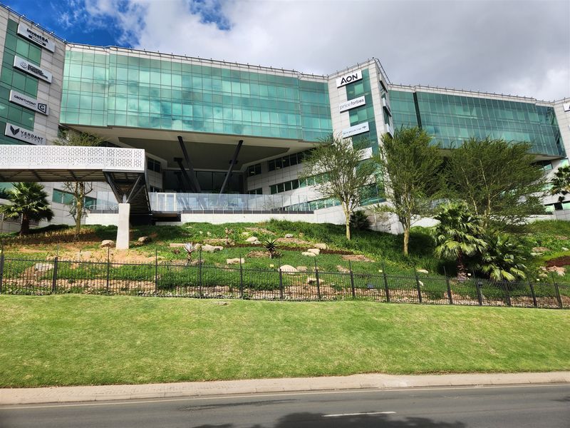 Modern multi-story office building featuring a glass curtain wall facade, elevated pedestrian access, and well-maintained landscaped grounds.