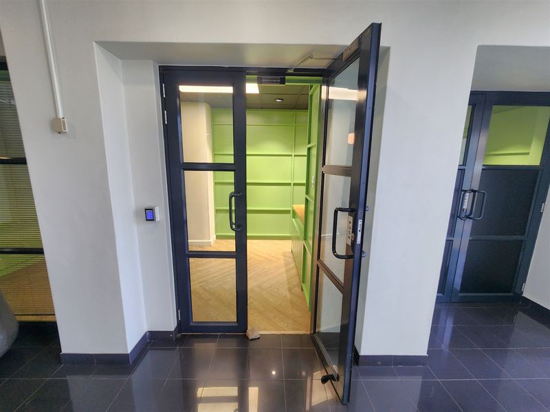 A modern office space featuring glass-panelled double doors, herringbone wood flooring, and distinctive green wall paneling, providing a bright and professional work environment.
