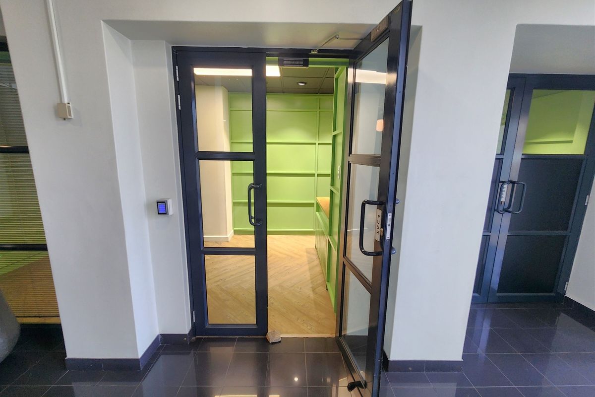 A modern office space featuring glass-panelled double doors, herringbone wood flooring, and distinctive green wall paneling, providing a bright and professional work environment.