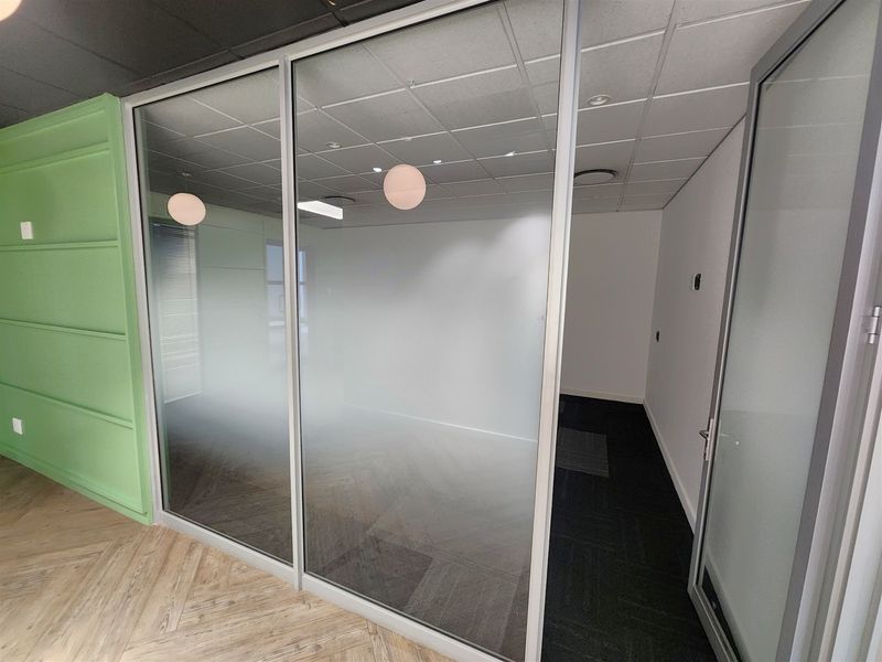 A modern, empty office space featuring a glass partition wall, suspended ceiling with recessed lighting, and a combination of herringbone and carpet flooring.