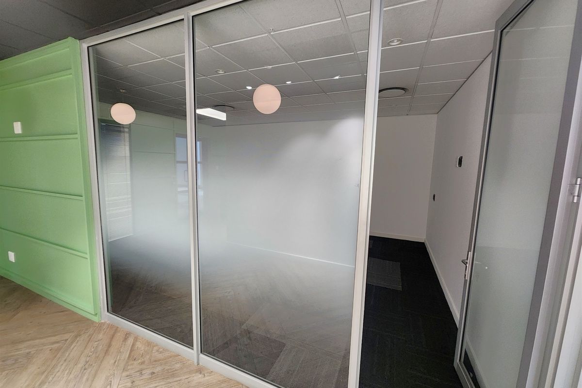 A modern, empty office space featuring a glass partition wall, suspended ceiling with recessed lighting, and a combination of herringbone and carpet flooring.
