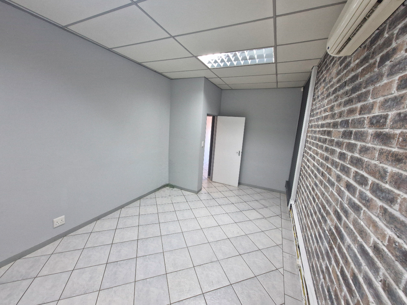 A vacant commercial office space featuring a modern exposed brick wall, tiled flooring, suspended ceiling with recessed lighting, and a wall-mounted air conditioning unit.