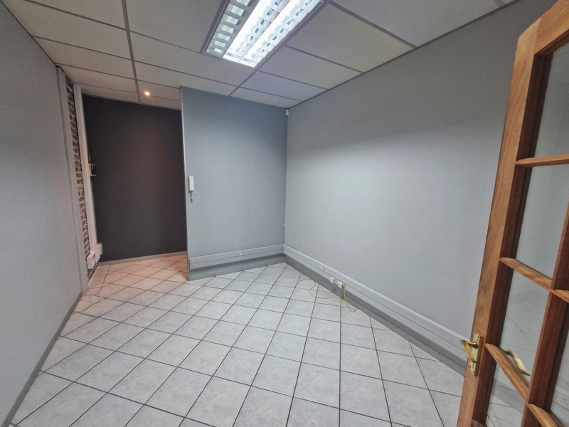A clean, empty office space featuring tiled flooring, suspended ceiling with integrated lighting, and neutral grey walls, suitable for a professional workspace.