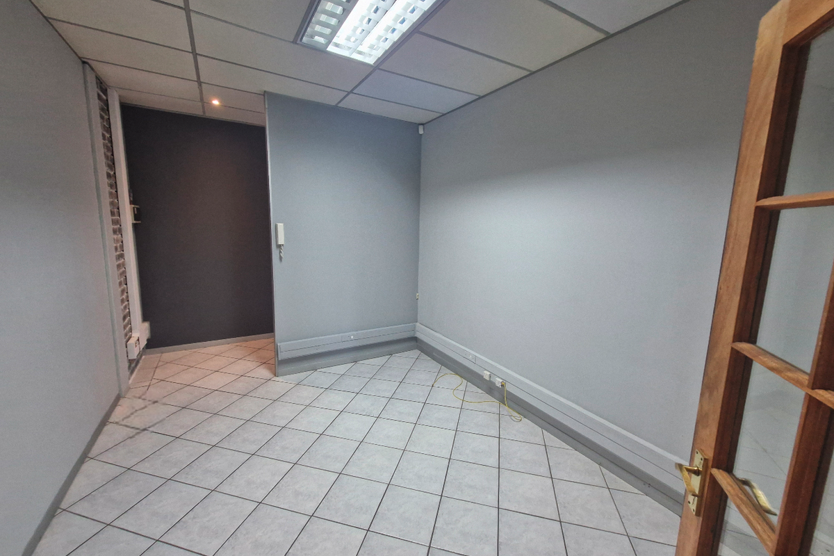 A clean, empty office space featuring tiled flooring, suspended ceiling with integrated lighting, and neutral grey walls, suitable for a professional workspace.