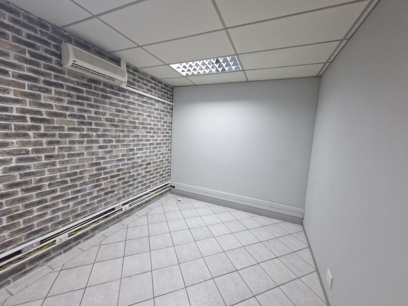 A vacant office space featuring a brick accent wall, tiled flooring, suspended ceiling with recessed lighting, and a wall-mounted air conditioning unit.
