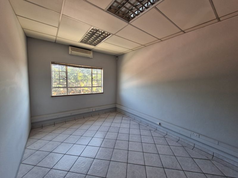 A vacant commercial office space featuring tiled flooring, a suspended ceiling with recessed lighting, and a wall-mounted air conditioning unit.