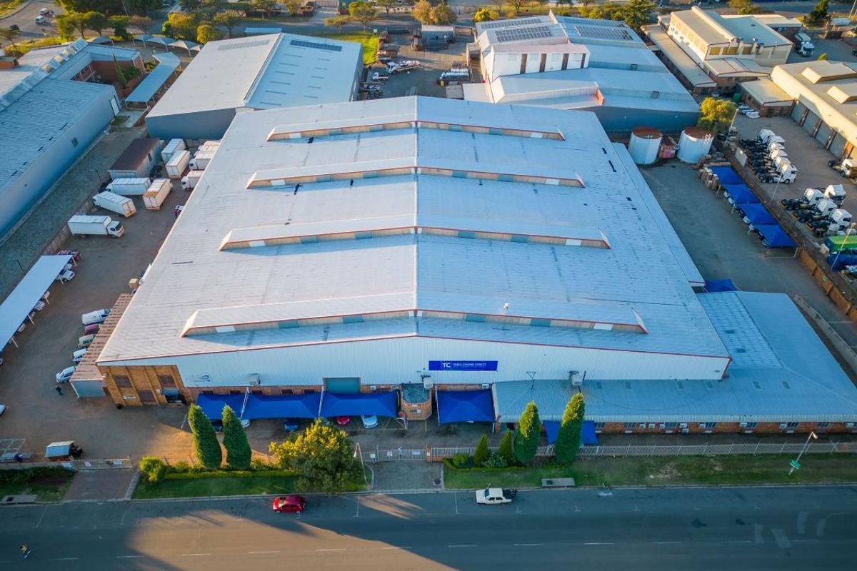 Aerial view of a large industrial warehouse facility featuring a metal roof with skylights, paved yard area, and perimeter security fencing.