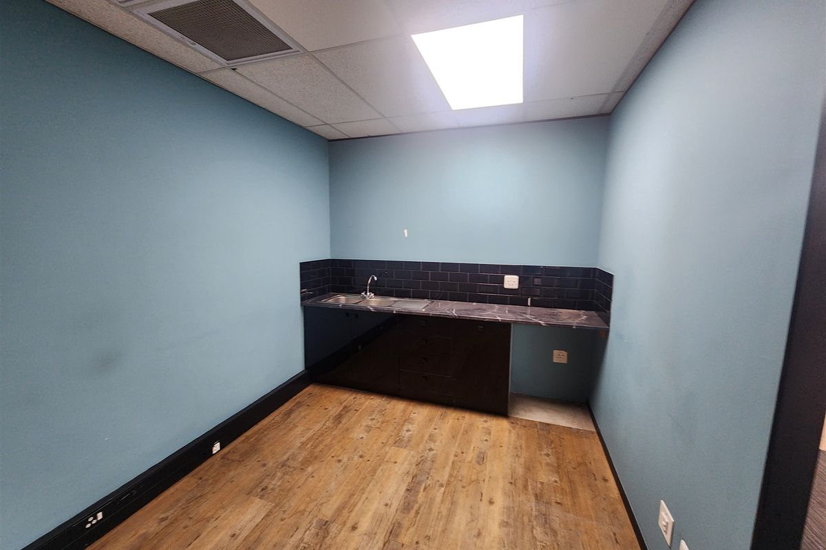 A clean, empty office kitchenette featuring dark cabinetry, a stainless steel sink, tiled backsplash, and wood-look laminate flooring.