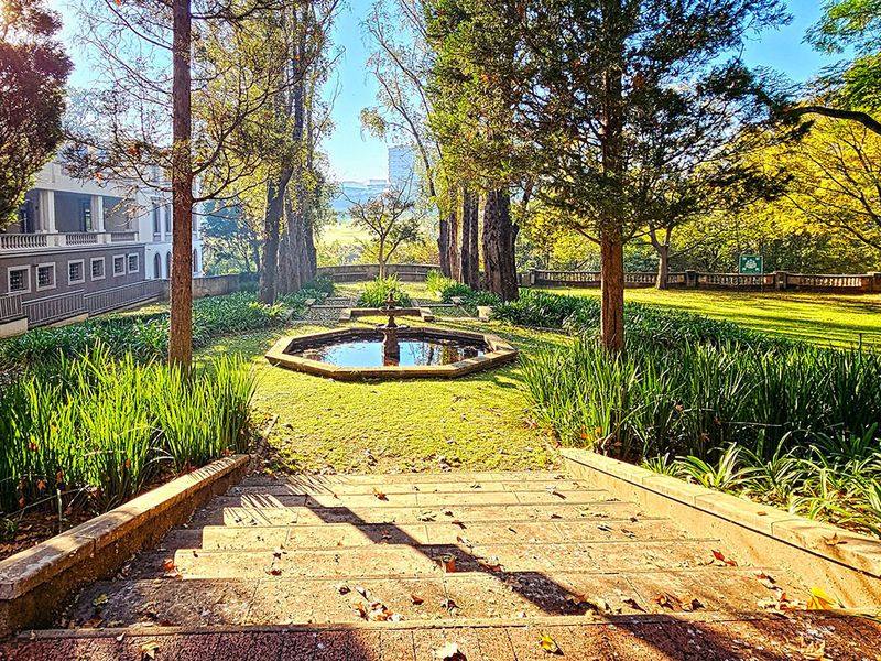A beautifully landscaped garden featuring a central stone fountain, mature trees, and stone stairs leading down to the lawn area.