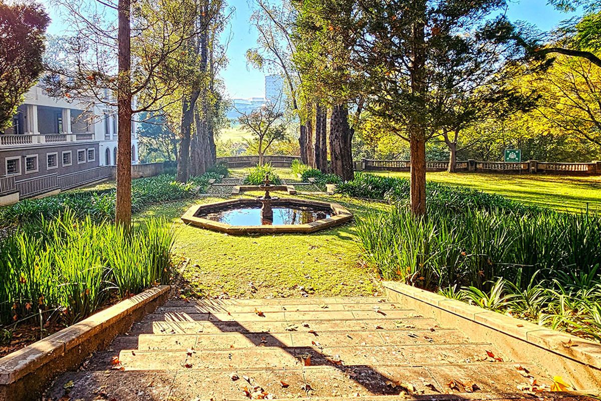 A beautifully landscaped garden featuring a central stone fountain, mature trees, and stone stairs leading down to the lawn area.