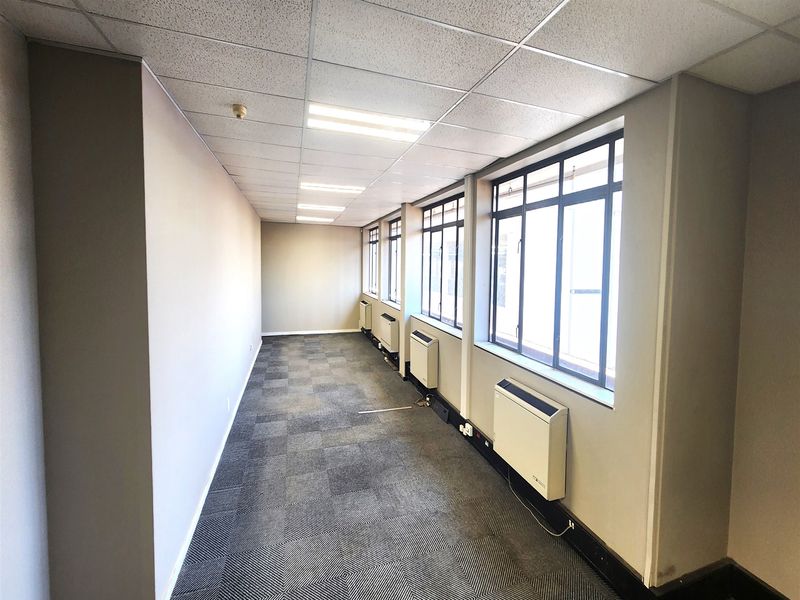 An empty, open-plan office space featuring carpeted flooring, suspended ceilings, large windows for natural light, and wall-mounted HVAC units.