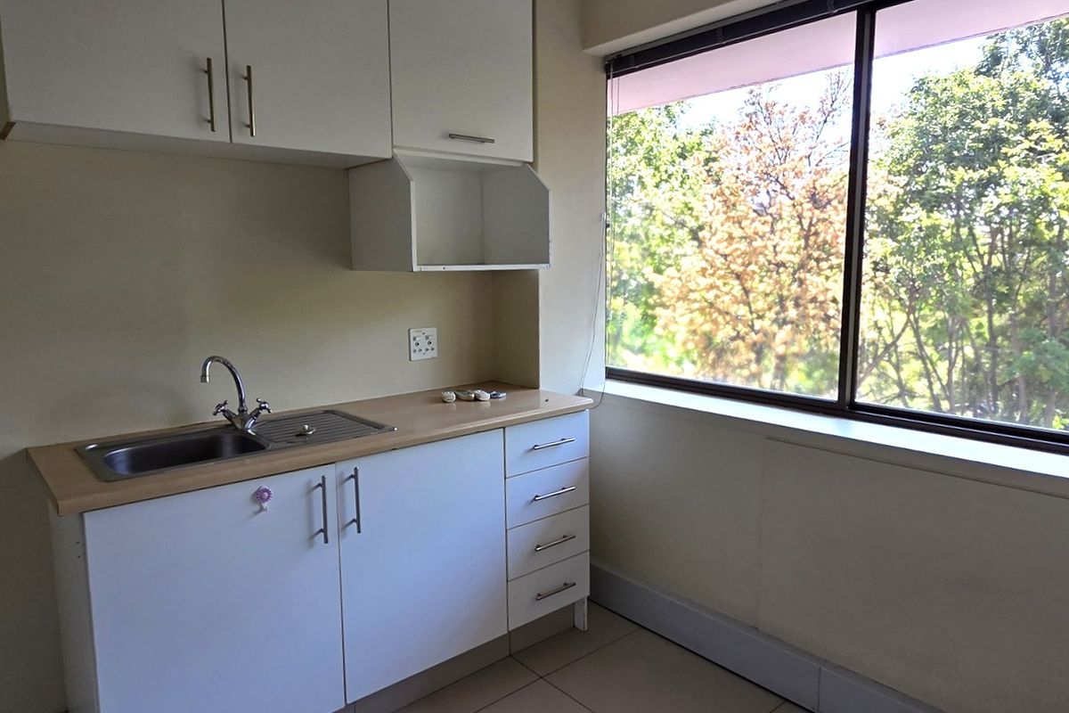 A compact office kitchenette featuring white built-in cabinetry, a stainless steel sink, and a large window providing natural light and a pleasant outdoor view.