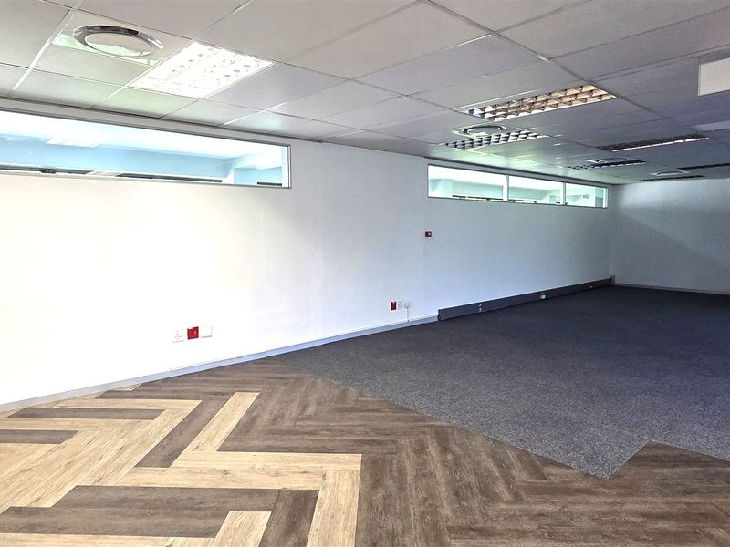 A spacious, vacant office area featuring a mix of herringbone vinyl and carpet flooring, suspended ceilings, and clerestory windows for natural light.