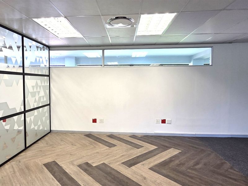A modern, empty office space featuring a glass partition wall, herringbone vinyl flooring, and a suspended ceiling with recessed lighting.