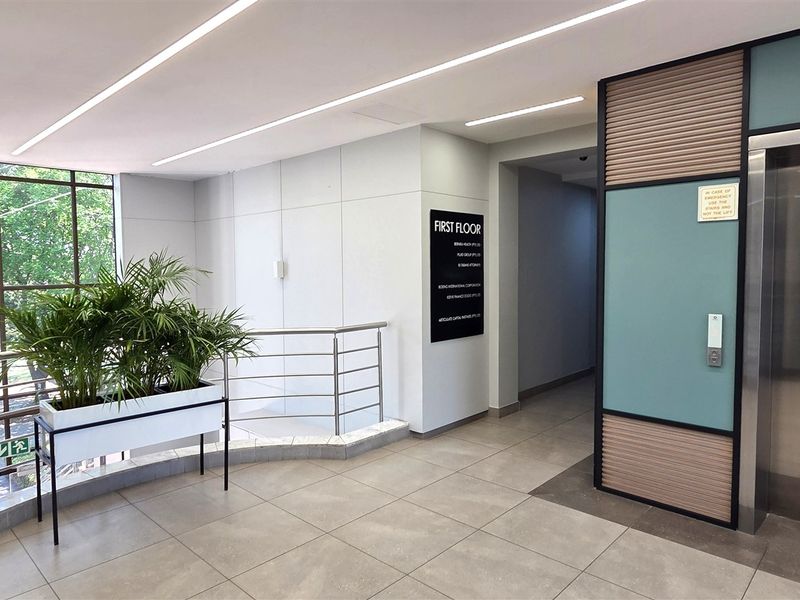 Modern office building lobby featuring a stainless steel elevator, large glass windows, clean tiled flooring, and professional linear lighting.