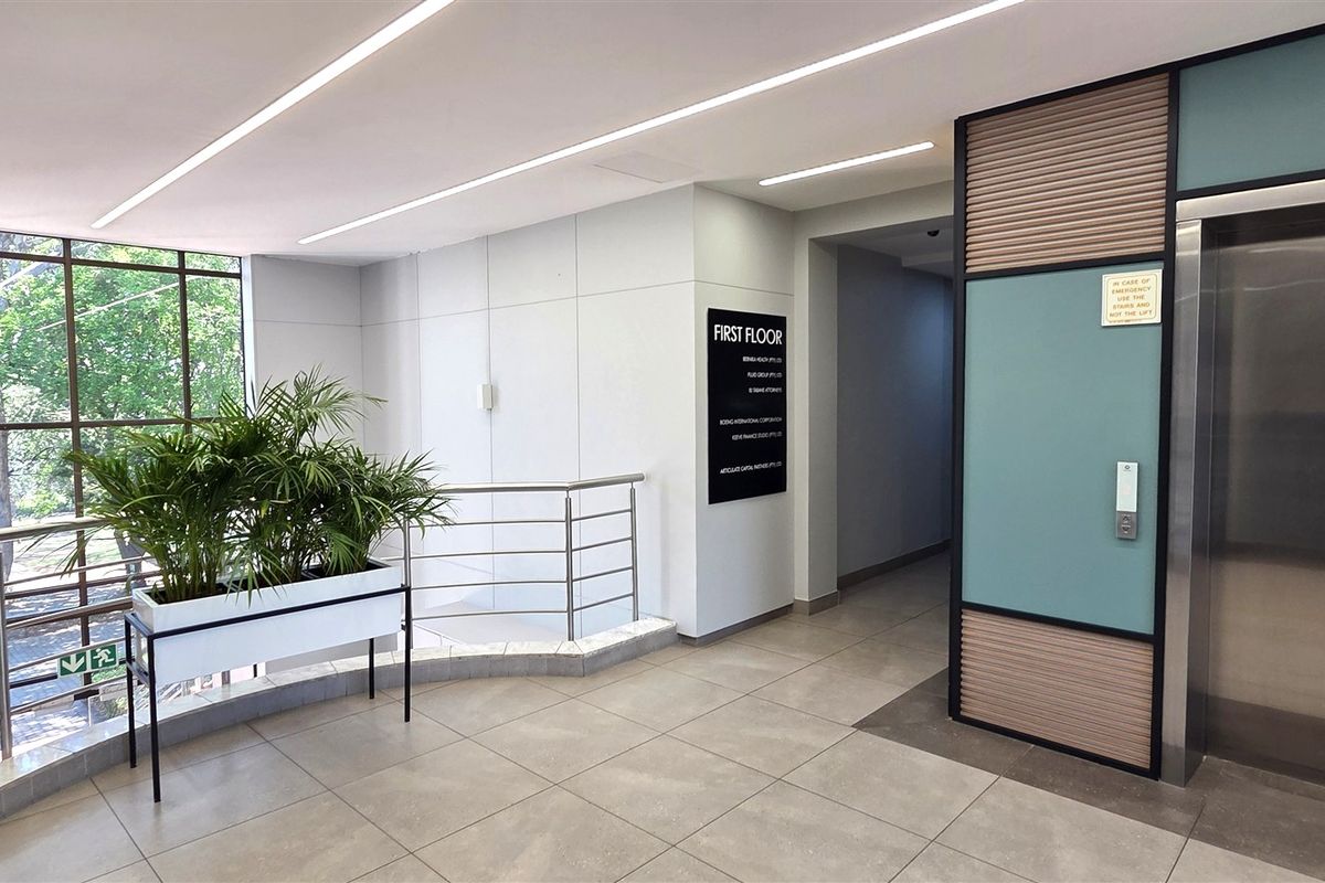Modern office building lobby featuring a stainless steel elevator, large glass windows, clean tiled flooring, and professional linear lighting.