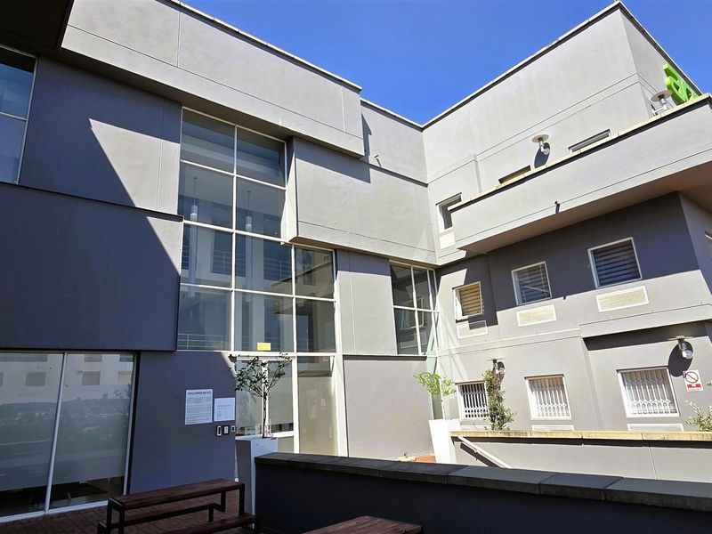 A modern office building exterior featuring a large glass facade, grey rendered walls, and a paved terrace area with integrated lighting.