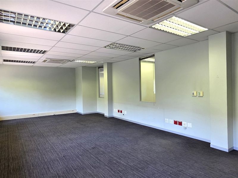 A spacious, empty office suite featuring a suspended ceiling with recessed lighting, ducted air conditioning, carpeted flooring, and glass partition elements.