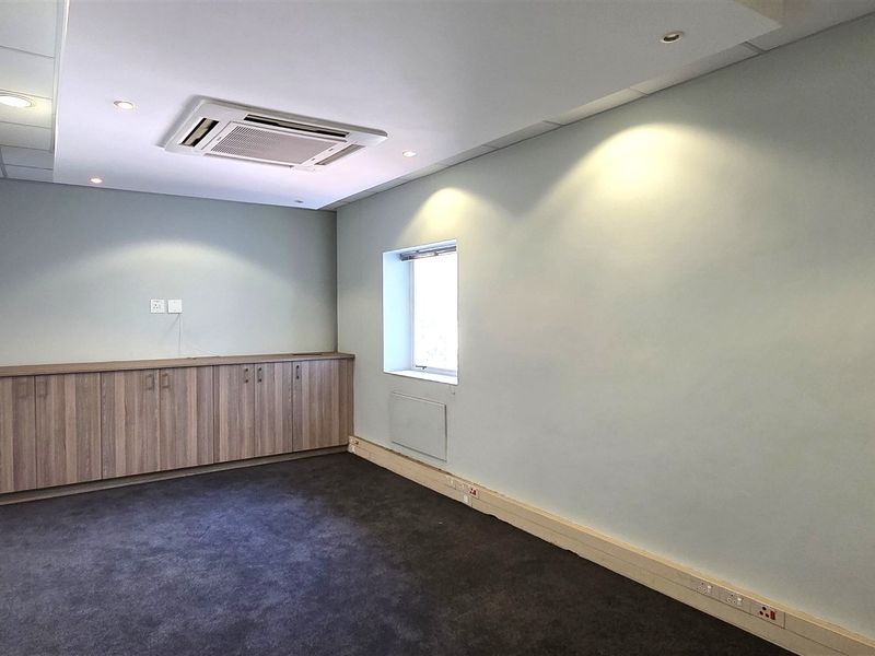 A clean, empty office space featuring built-in wooden cabinetry, recessed ceiling lighting, a ceiling-mounted air conditioning unit, and dark carpeted flooring.