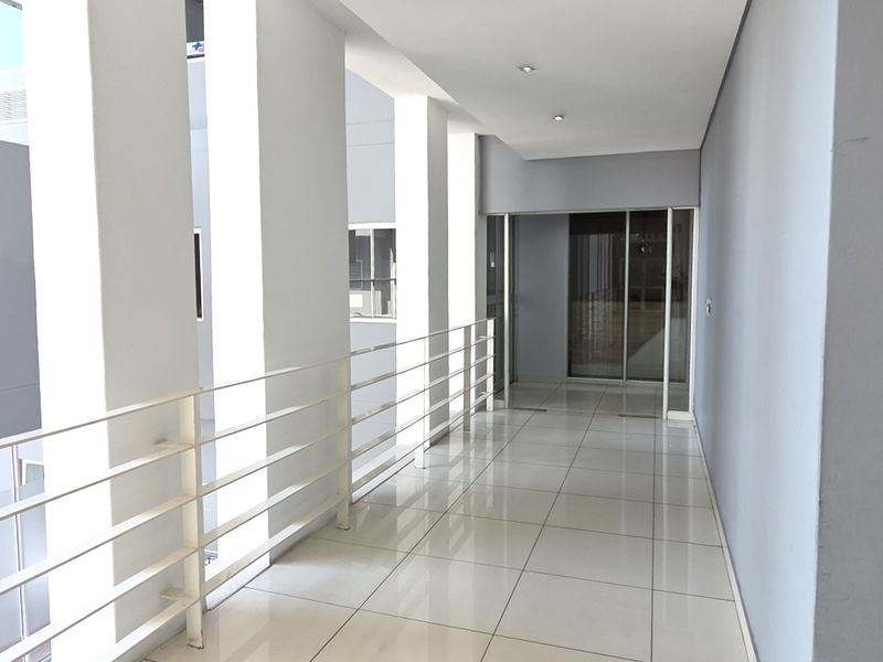 A bright, modern office hallway featuring polished tiled flooring, a white metal balustrade, and recessed ceiling lighting leading to a glass-paneled entrance.