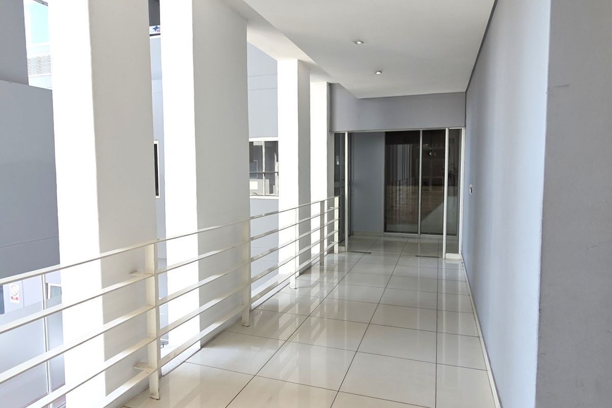 A bright, modern office hallway featuring polished tiled flooring, a white metal balustrade, and recessed ceiling lighting leading to a glass-paneled entrance.