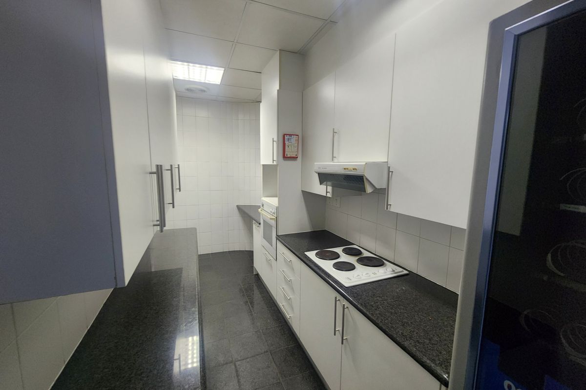 A clean, functional office kitchenette featuring white cabinetry, dark granite countertops, an electric hob, built-in oven, and tiled finishes.