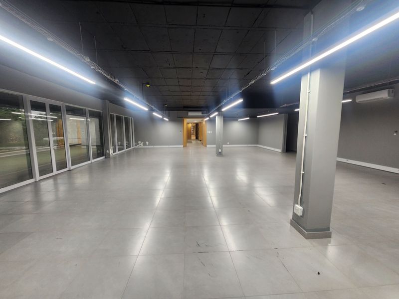 Spacious open-plan office area featuring modern tiled floors, exposed industrial-style ceiling, linear lighting, and large glass doors providing ample natural light.