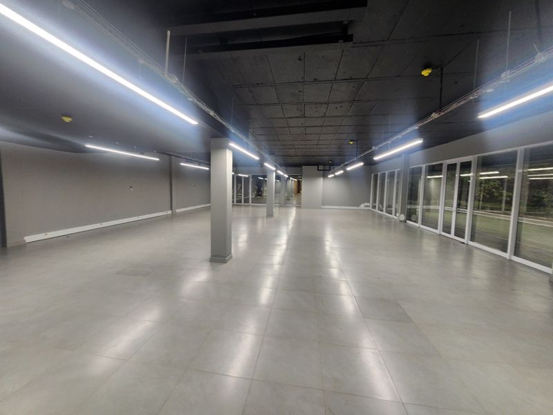 A spacious, modern open-plan office area featuring polished tiled floors, exposed concrete ceilings, linear lighting, and extensive floor-to-ceiling glass windows.