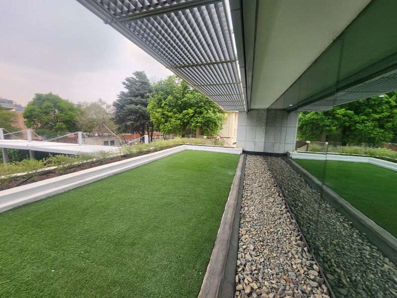 Modern elevated balcony featuring artificial turf, a gravel drainage border, and a sleek metal louvered canopy, adjacent to a full-height glass office facade.
