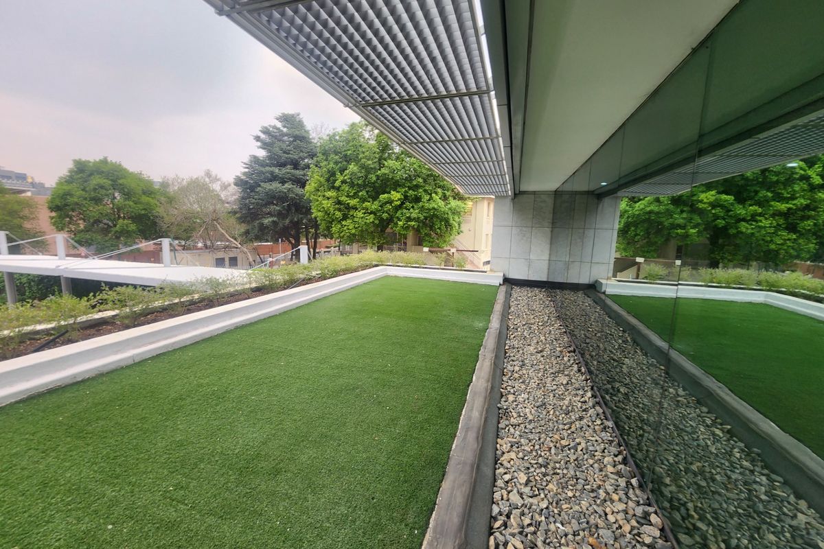 Modern elevated balcony featuring artificial turf, a gravel drainage border, and a sleek metal louvered canopy, adjacent to a full-height glass office facade.