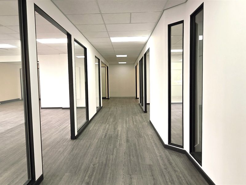 A modern, well-lit office hallway featuring glass partition walls, suspended ceilings, and durable laminate flooring, providing a clean and professional workspace aesthetic.