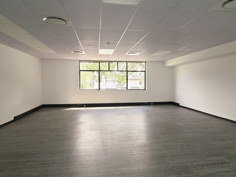 A spacious, empty office area featuring a modern suspended ceiling, recessed lighting, and dark laminate flooring with ample natural light from a large window.