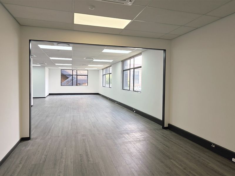 A spacious, open-plan office area featuring modern laminate flooring, recessed ceiling lighting, and large windows providing ample natural light.