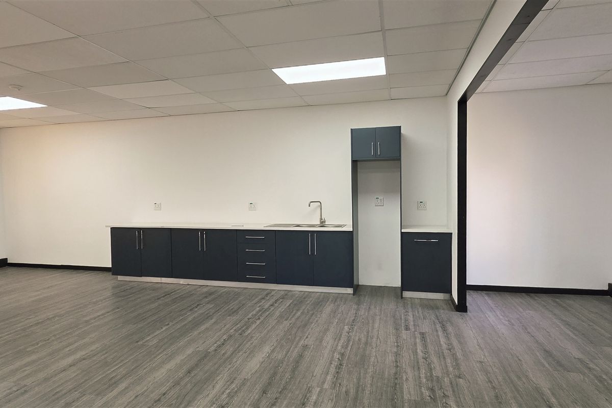 A clean, modern office kitchenette featuring dark blue cabinetry, a stainless steel sink, and wood-look laminate flooring in an open-plan workspace.