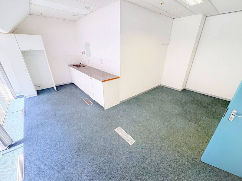 An empty office space featuring a small built-in kitchenette with a sink, suspended ceiling tiles, and carpeted flooring.