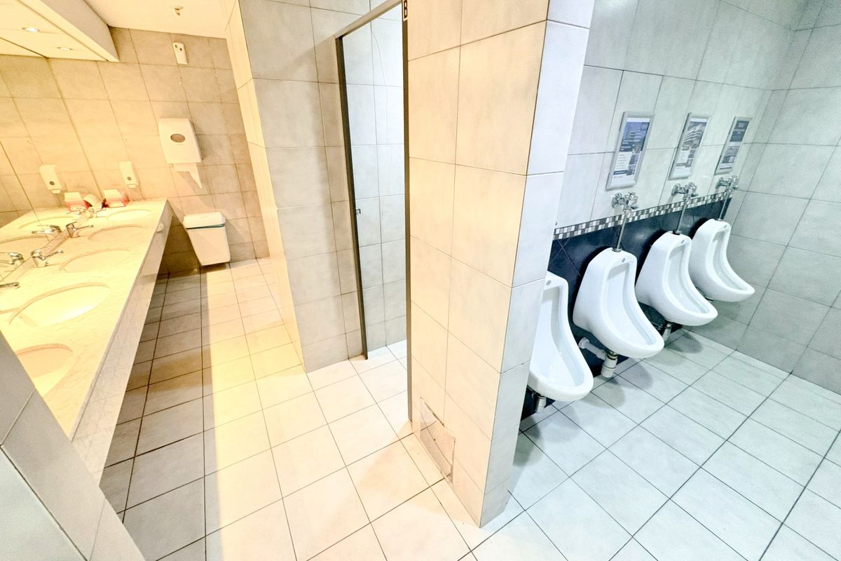 A clean, modern commercial office bathroom featuring multiple wall-mounted urinals, a long vanity countertop with integrated sinks, and neutral floor-to-ceiling tiling.