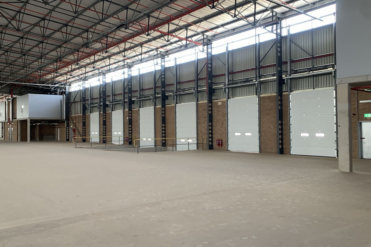 Spacious industrial warehouse featuring high ceilings, exposed steel trusses, multiple roller shutter doors, and polished concrete flooring, suitable for logistics or manufacturing operations.