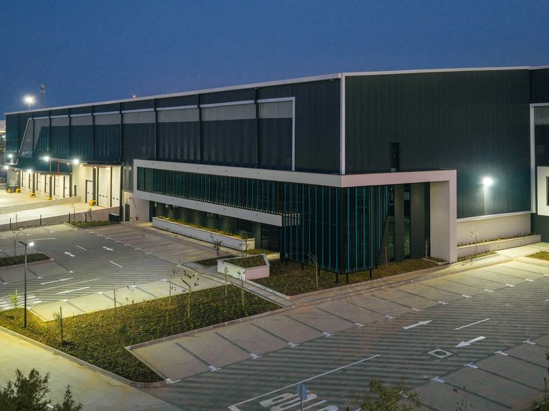 Modern industrial facility featuring a large warehouse structure with integrated loading docks, extensive paved parking, and professional exterior lighting at dusk.