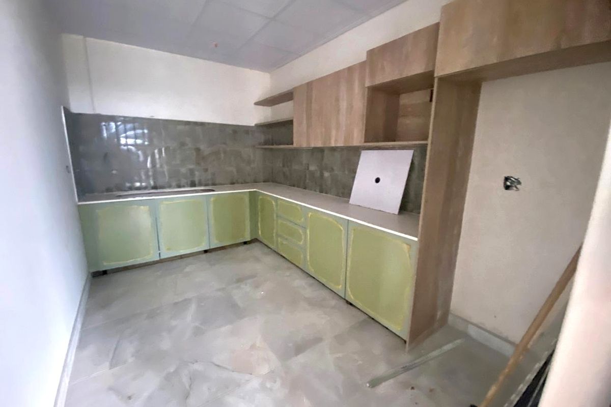 An unfinished kitchen area featuring light green base cabinetry, wood-tone upper shelving, tiled backsplash, and a drop-in sink, awaiting final installation and finishing touches.
