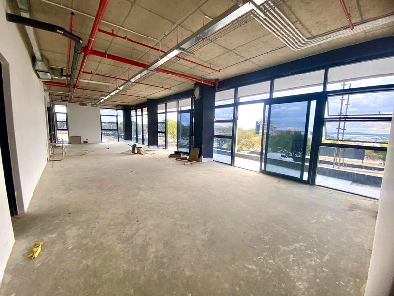 Spacious open-plan industrial office area featuring exposed concrete ceilings, fire sprinkler systems, and expansive floor-to-ceiling windows with natural light.