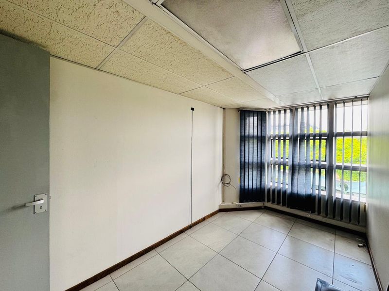 An empty office space featuring tiled flooring, a suspended ceiling, and large windows with vertical blinds, offering a blank canvas for professional use.