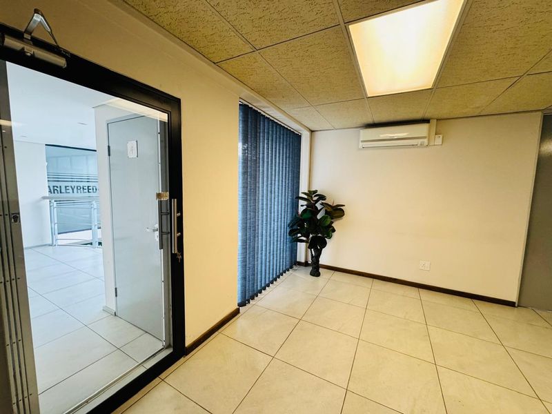 A bright, empty office space featuring tiled flooring, a suspended ceiling with recessed lighting, and a wall-mounted air conditioning unit.