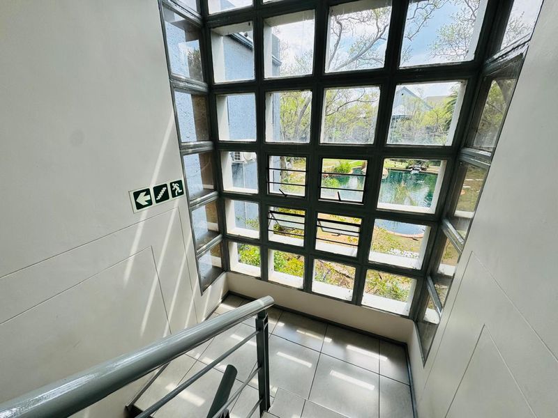 A bright stairwell landing featuring a large grid-pattern window overlooking the garden and pool area, with tiled flooring and a metal handrail.