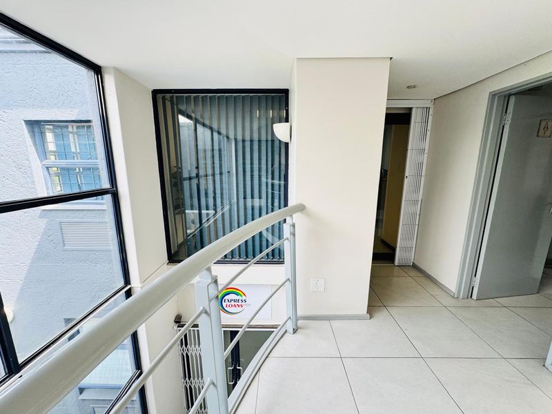 A bright, tiled office hallway featuring a metal balustrade, large windows with vertical blinds, and a folding security gate for access control.