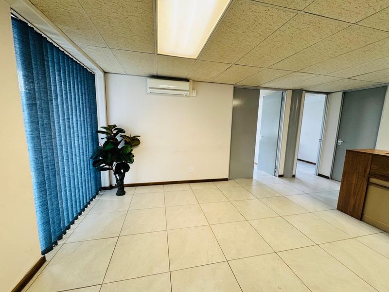 A bright, open-plan office space featuring tiled flooring, a drop ceiling with integrated lighting, and a wall-mounted air conditioning unit.