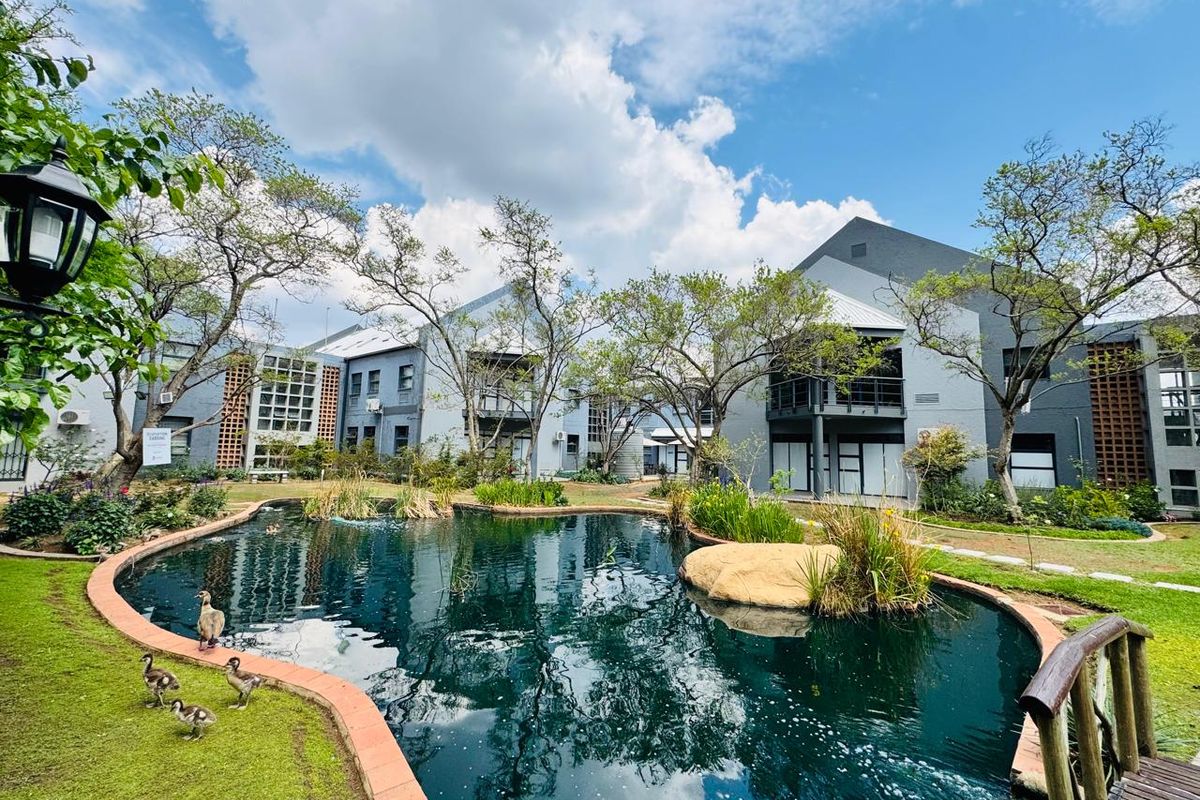 A scenic view of the office complex featuring a landscaped pond, wooden bridge, and well-maintained multi-story building exteriors with balconies.