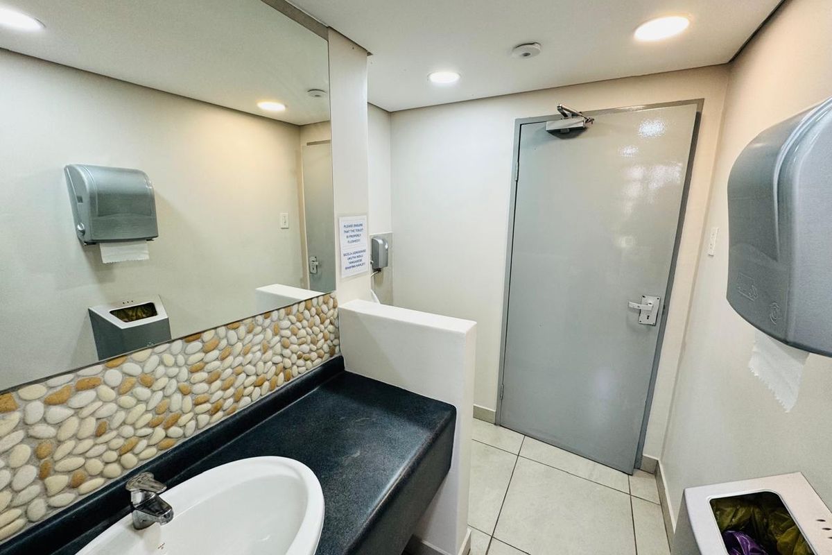 A clean and functional office bathroom featuring a vanity basin, large wall mirror, tiled splashback, and durable tiled flooring.