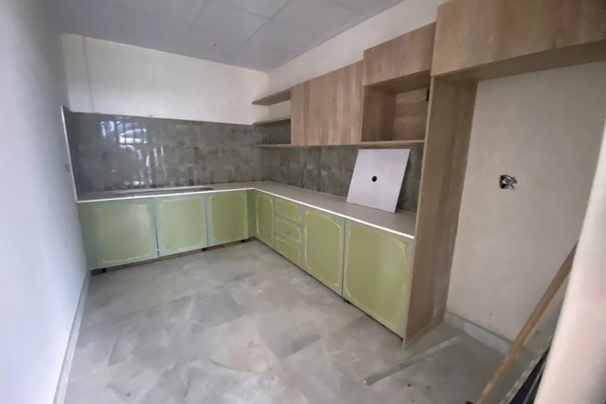 An unfinished kitchen area featuring light green base cabinetry, wood-tone upper cabinets, tiled backsplash, and neutral floor tiles, currently awaiting final installation and hardware.