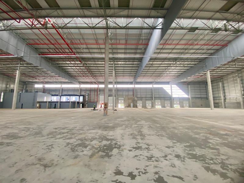 Expansive industrial warehouse space featuring high ceilings with exposed steel trusses, fire sprinkler systems, and multiple roller shutter doors for logistics access.