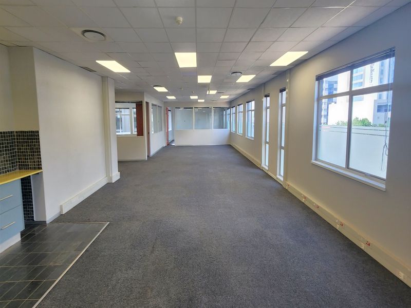 Spacious open-plan office area featuring large windows, suspended ceilings with recessed lighting, and a small kitchenette corner.
